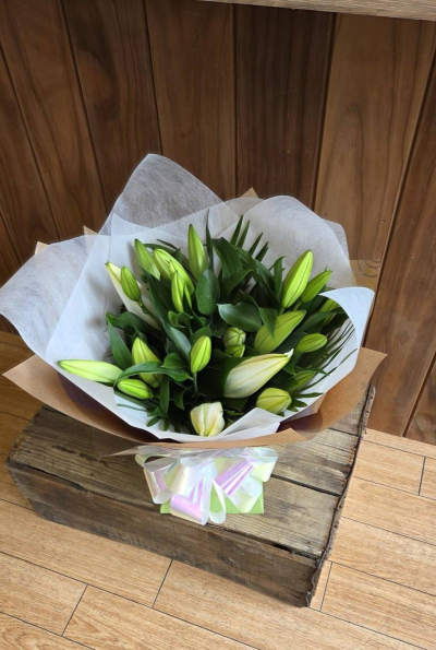 Lily Handtied - A graceful hand-tied bouquet of elegant white lillies nestled among rich fresh greenery and wrapped in soft neutral tones. Beautifully presented with a delicate finishing bow, this timeless arrangement offers a sophisticated and serene gift suitable for any special occasion.